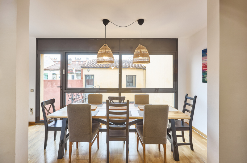 Dining room with a long wooden table and eight chairs, large glass doors showing an urban exterior, two woven pendant lights overhead.
