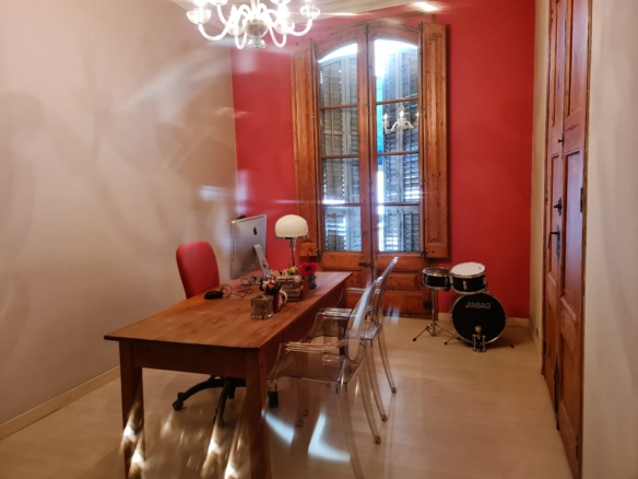 Home office with wooden desk and iMac, red chair, clear acrylic chairs, white lamp; red wall and large wooden window shutters; drum set in the corner.