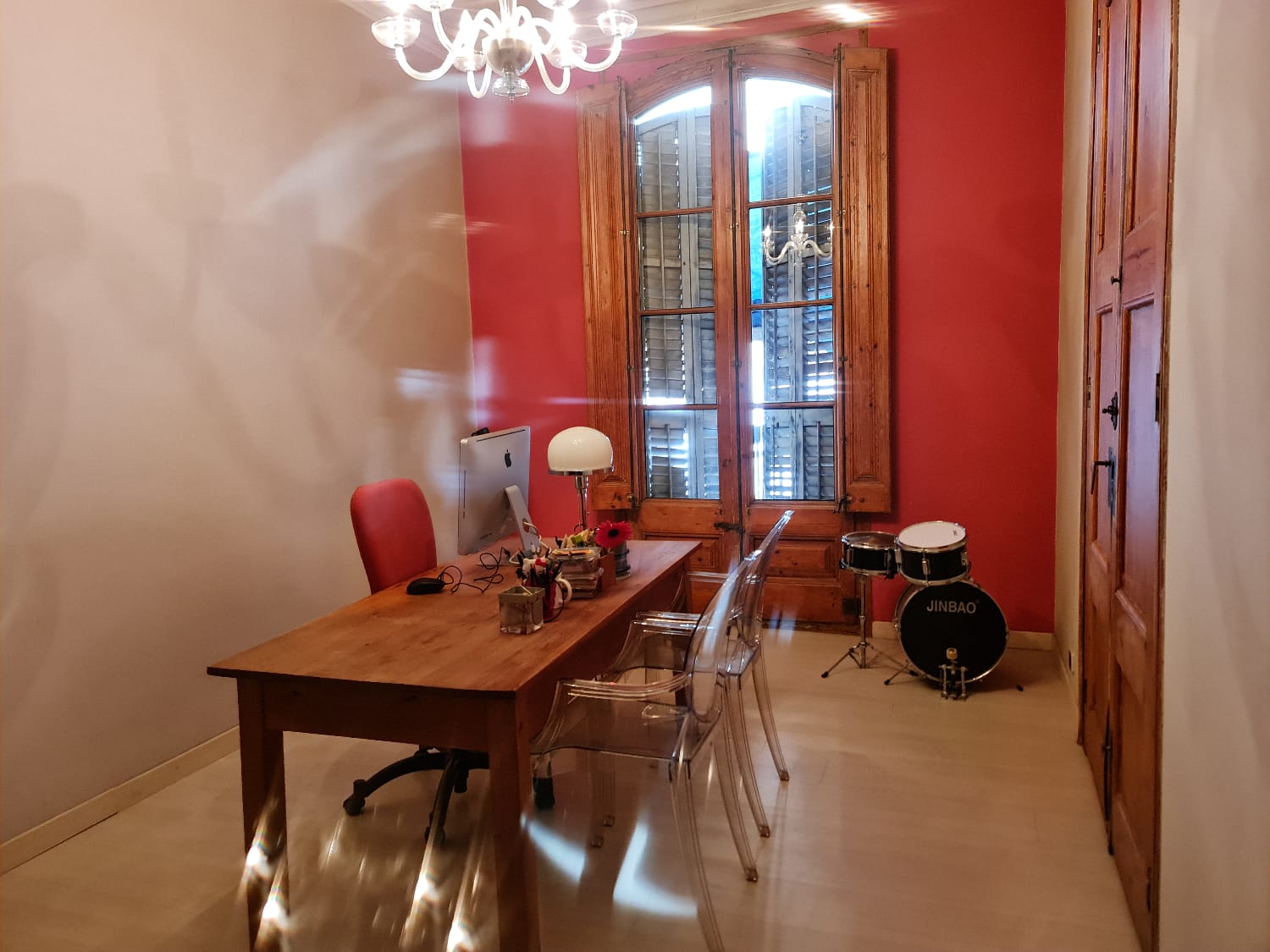 Home office with wooden desk and iMac, red chair, clear acrylic chairs, white lamp; red wall and large wooden window shutters; drum set in the corner.