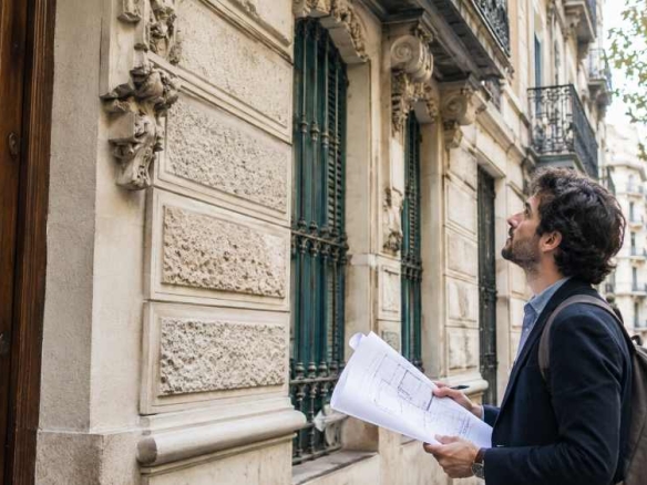 A man in a dark blazer with a backpack reads architectural blueprints outside a historic stone building on a city street.