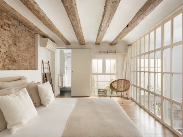 Bright bedroom with exposed wooden beams, brick accent wall, and a glass-walled area on the right; bed with beige and white linens.