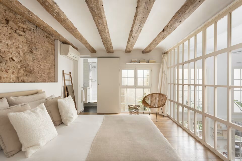 Bright bedroom with exposed wooden beams, brick accent wall, and a glass-walled area on the right; bed with beige and white linens.