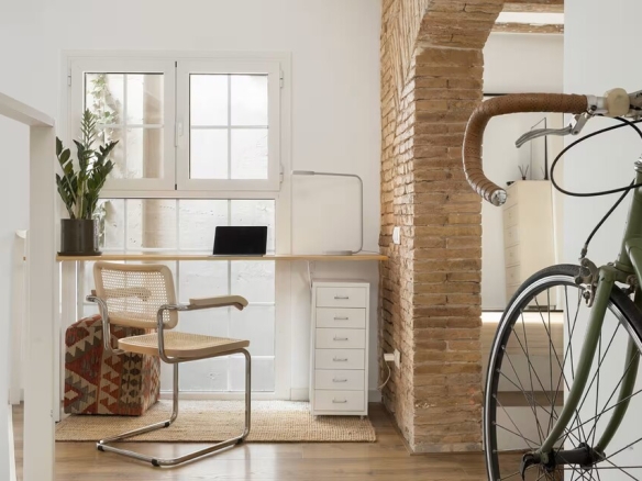 Bright home office by a window: a wooden desk with a laptop, wicker chair, potted plant, and a green bicycle beside a brick pillar.
