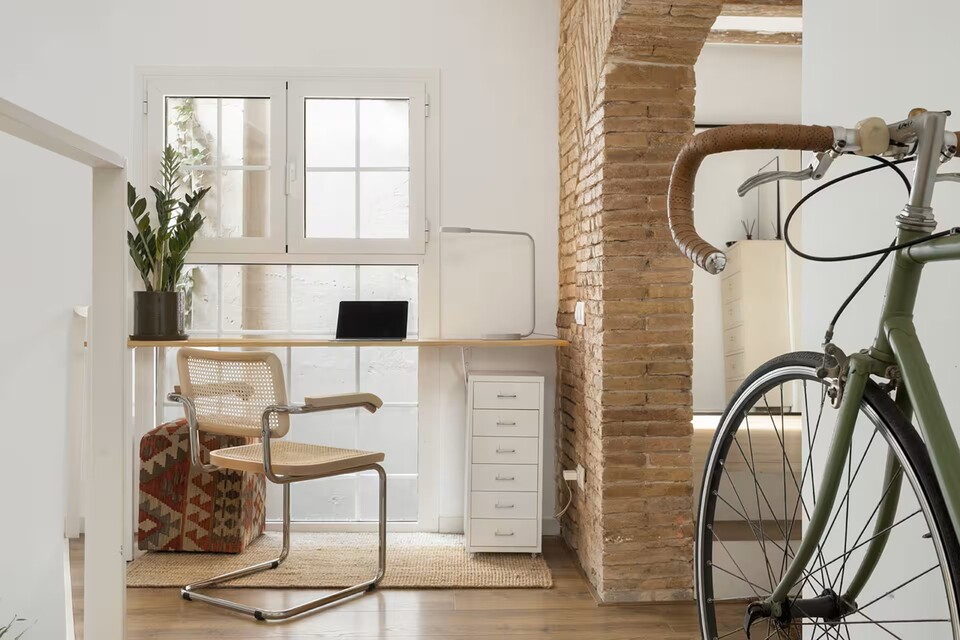 Bright home office by a window: a wooden desk with a laptop, wicker chair, potted plant, and a green bicycle beside a brick pillar.