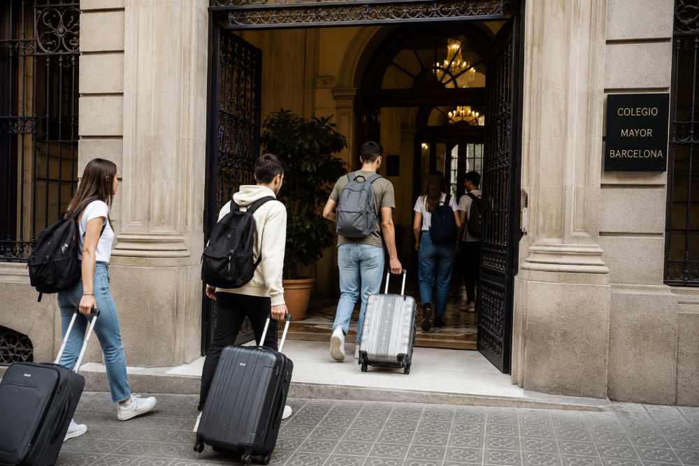 Group of students with rolling suitcases entering a historic campus building in Barcelona - Tu inmobiliaria de confianza en Barcelona Group of students with rolling suitcases entering a historic campus building in Barcelona