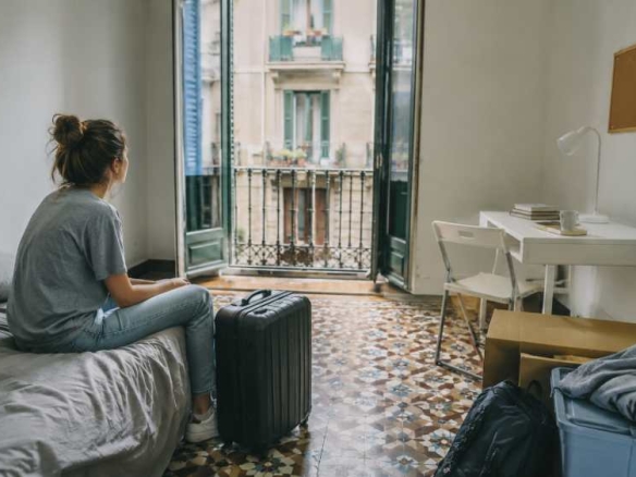 Person sits on a bed in a cluttered room, looking toward an open balcony door with a suitcase beside them.