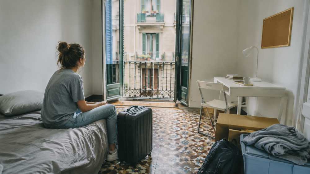 Person sits on a bed in a cluttered room, looking toward an open balcony door with a suitcase beside them.