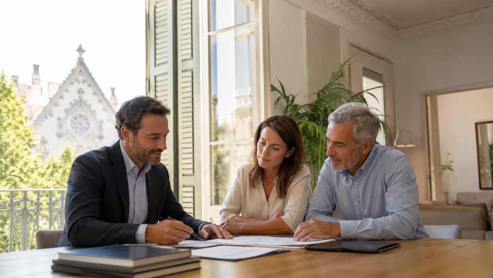 Three professionals discuss documents at a wooden table in a bright office, with a large window and greenery outside.