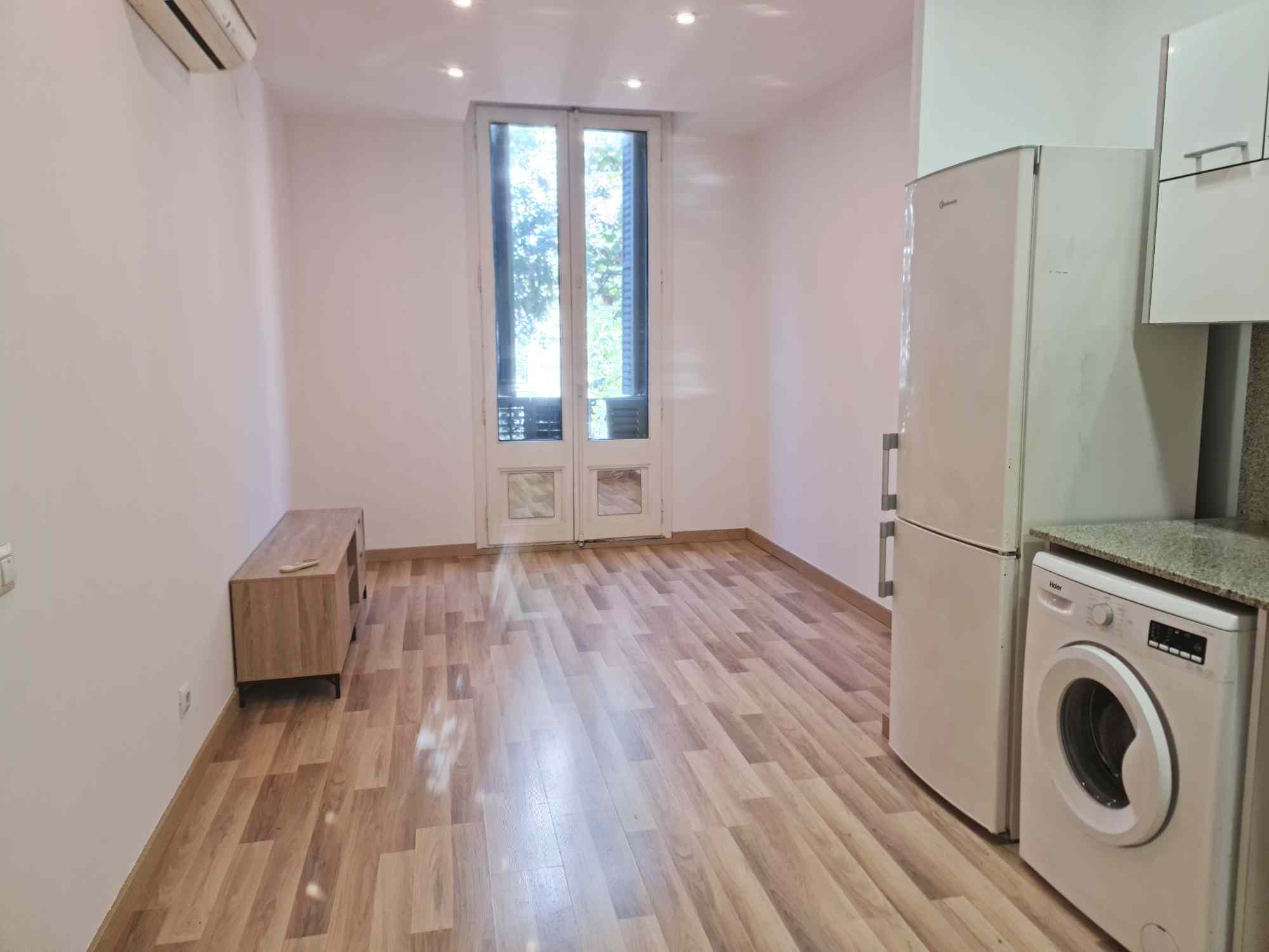 Narrow kitchen with light wood floor, white walls, and a tall white refrigerator on the right; washing machine beneath the counter and a windowed door at the far end.