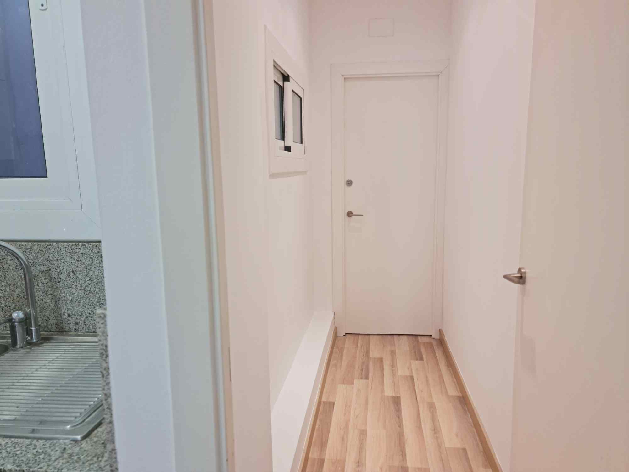 White hallway with light wood floors, a closed white door at the end, and a kitchen counter with a window on the left.