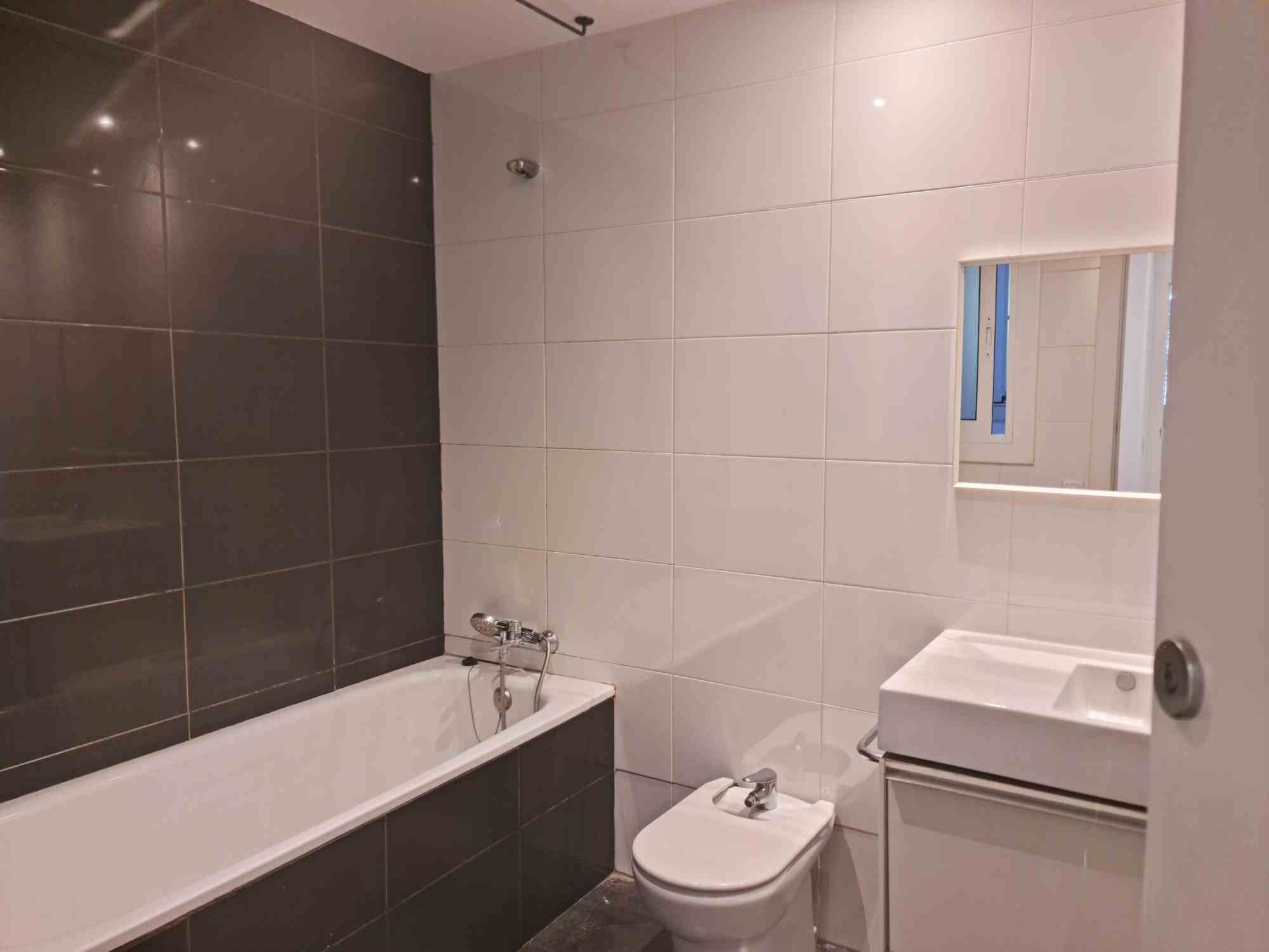 Modern bathroom with white wall tiles and a dark tile bathtub surround, featuring a bathtub, a toilet and bidet, and a wall mirror above a sink.