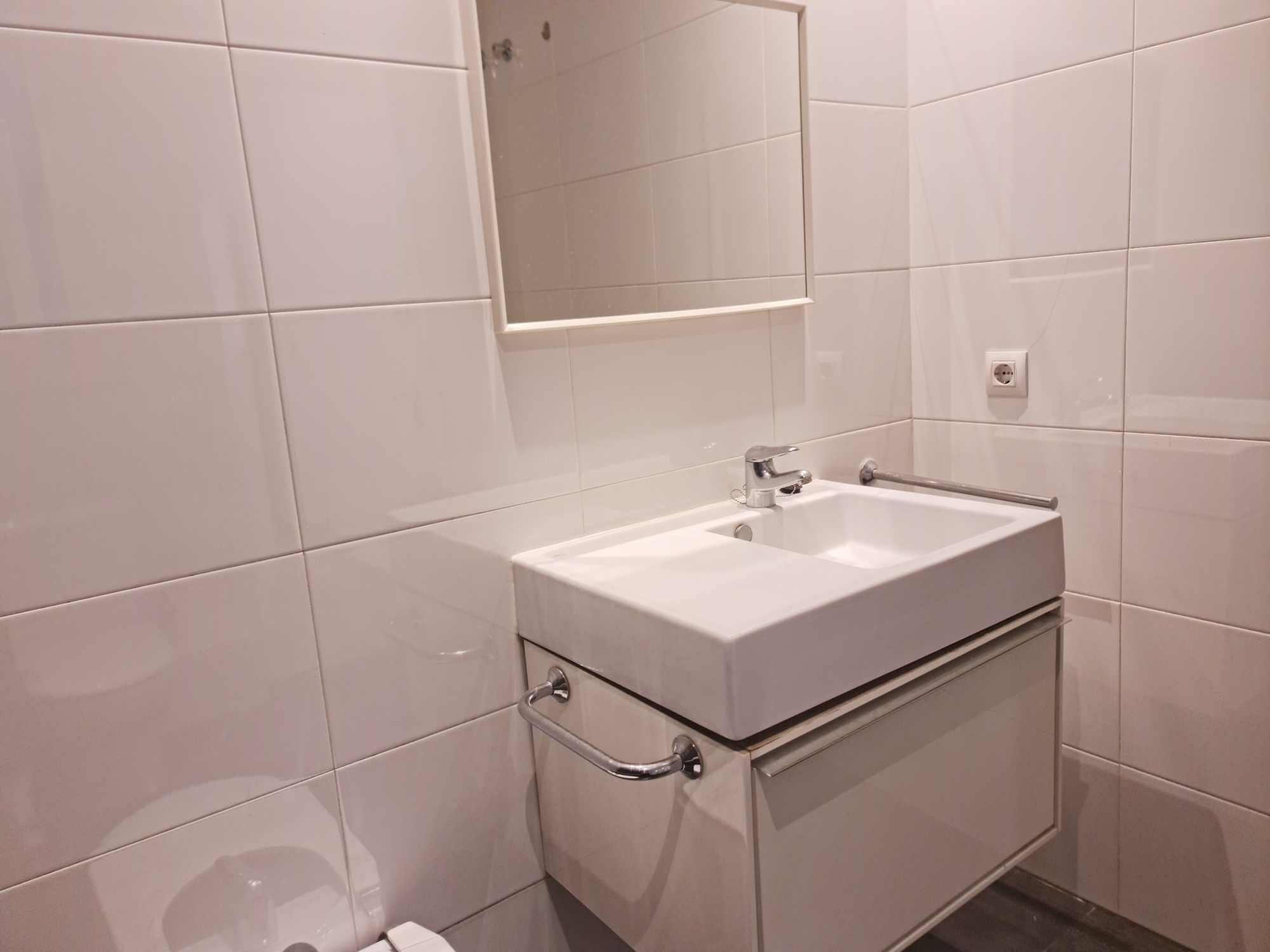 White tiled bathroom featuring a wall-mounted rectangular sink, chrome faucet, and a small mirror above. A towel rail runs along the side wall and a nearby electrical outlet is visible.