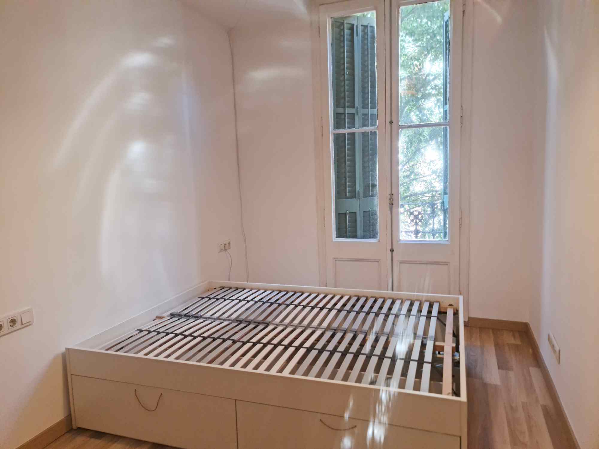 Empty white bedroom with a low-profile bed frame showing wooden slats and built-in drawers, near a windowed door and hardwood floor.