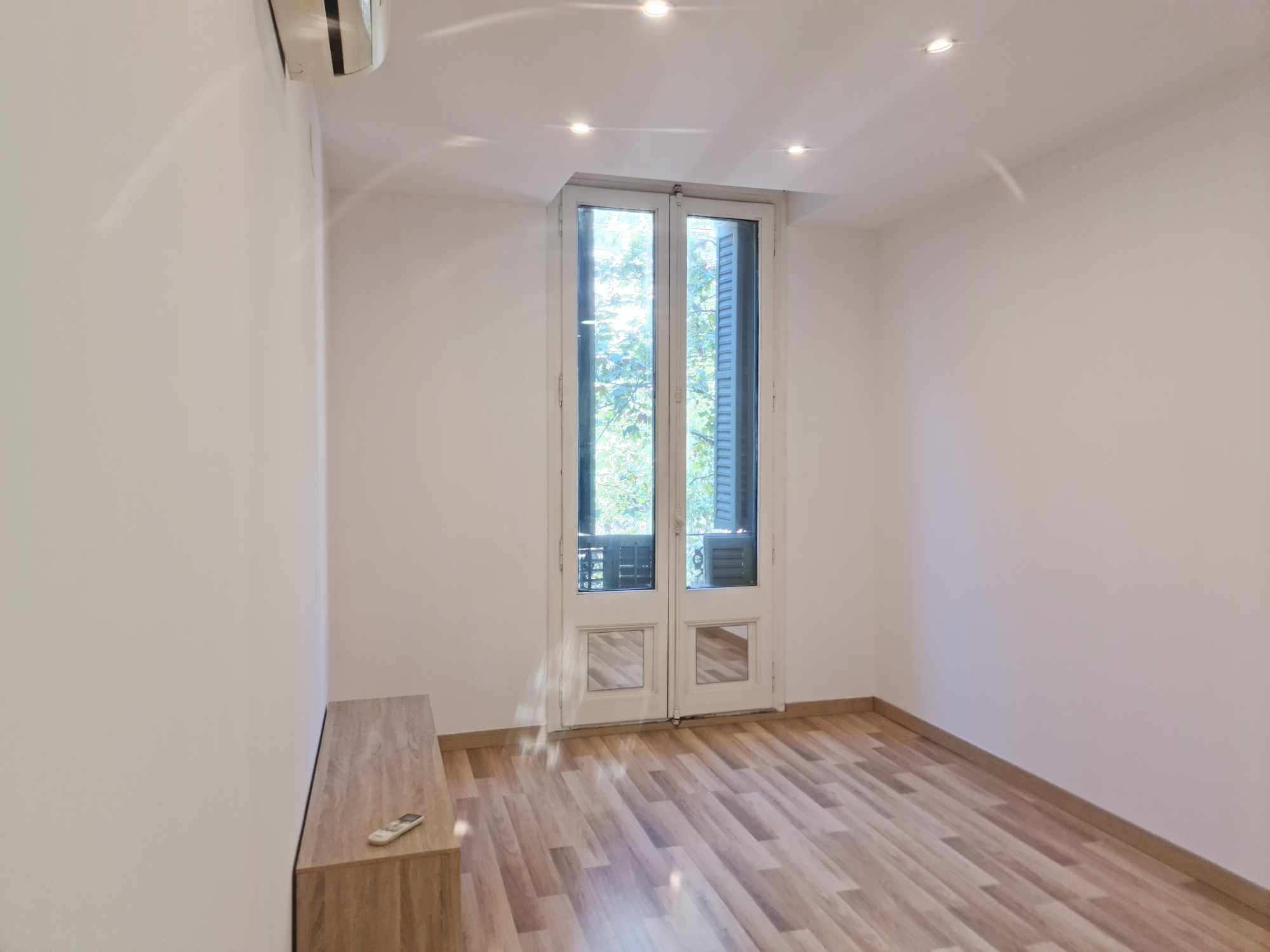 Narrow white-walled room with a tall glazed French door to a small outdoor balcony, wooden plank floor, and a built-in wooden bench with a remote on it.