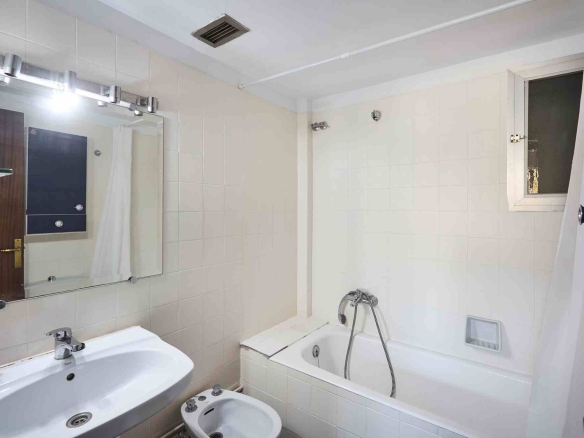 Small white-tiled bathroom featuring a bathtub with handheld shower, a sink and bidet, a wall mirror, and a window with a white frame.
