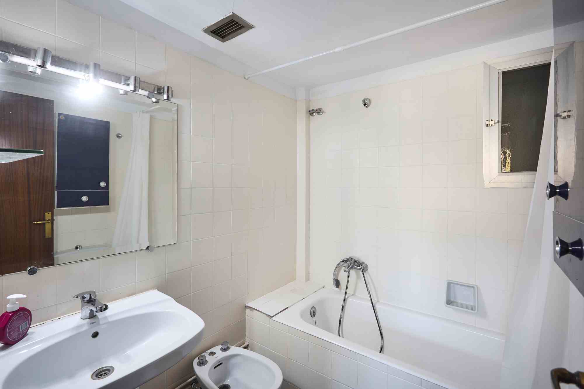 Small white-tiled bathroom featuring a bathtub with handheld shower, a sink and bidet, a wall mirror, and a window with a white frame.
