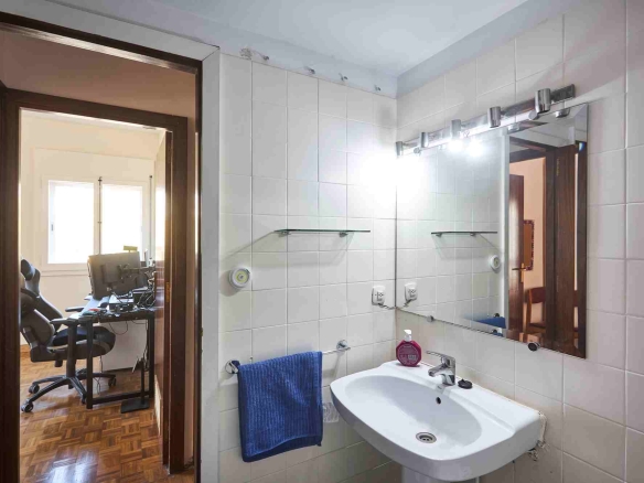 Bathroom with white tile walls, a sink and large mirror; a blue towel hangs on a rail, doorway to a home office with a computer desk visible beyond.