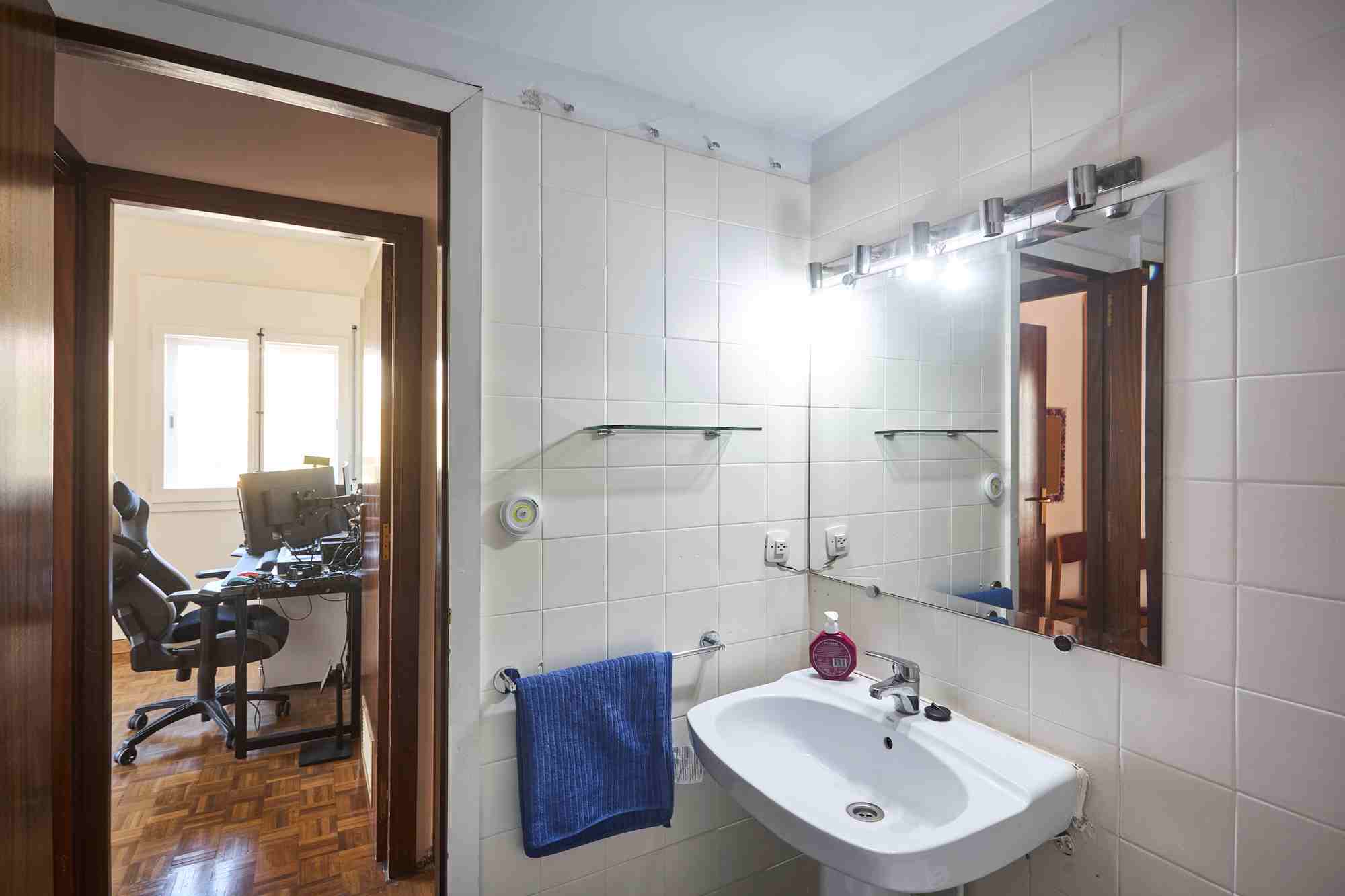 Bathroom with white tile walls, a sink and large mirror; a blue towel hangs on a rail, doorway to a home office with a computer desk visible beyond.