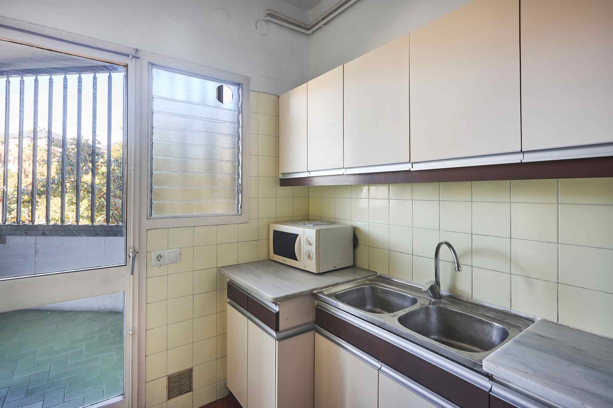 Small kitchen with cream cabinets, beige tiled backsplash, a double sink, and a microwave on the counter as part of the workspace outside a door window.