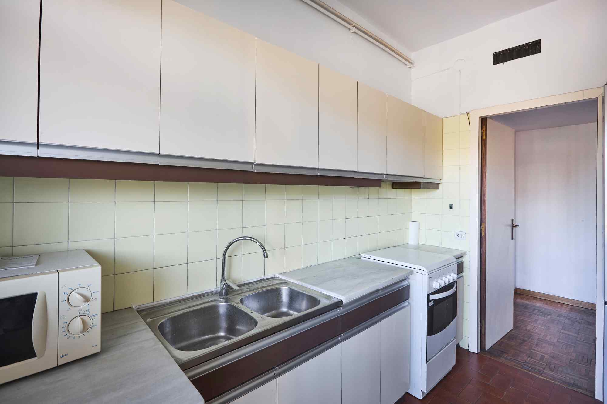 Kitchen with row of white wall cabinets above a pale tiled backsplash, stainless double sink, and white appliances along a marble countertop, doorway to adjacent room visible.