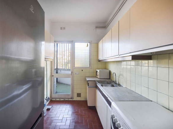 Narrow galley kitchen with white cabinets, cream tile walls, a stainless fridge on the left, and a window at the far end; microwave and sink along the right.