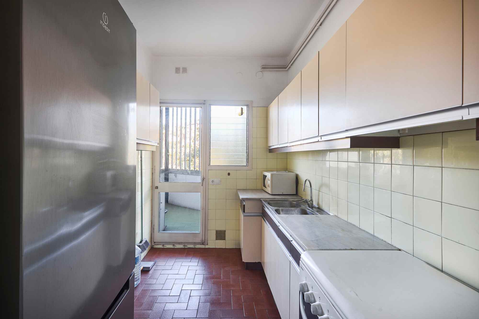 Narrow galley kitchen with white cabinets, cream tile walls, a stainless fridge on the left, and a window at the far end; microwave and sink along the right.