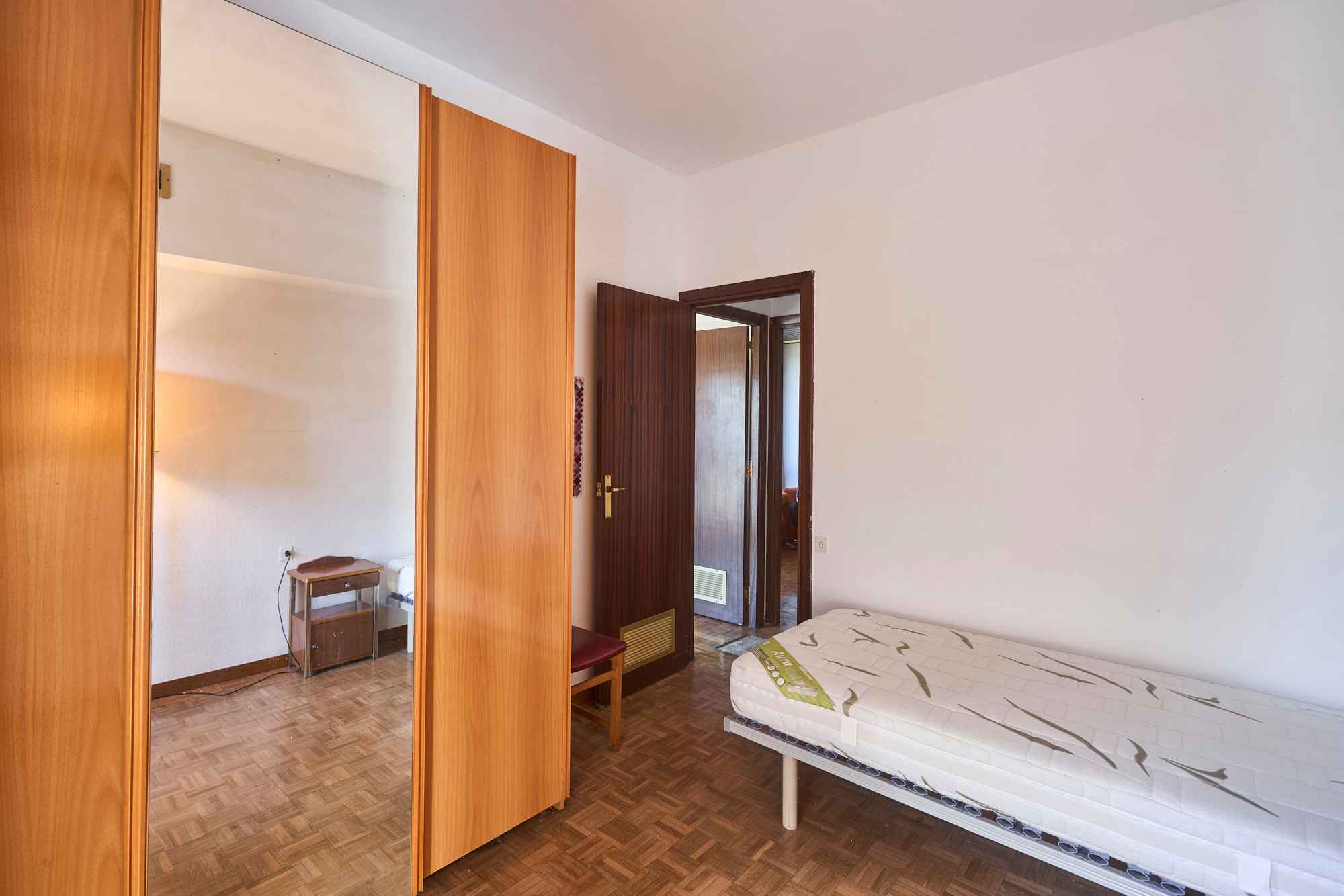 Bedroom with tall wooden wardrobes and a mirrored door, a bed at the edge, and an open doorway to another room.