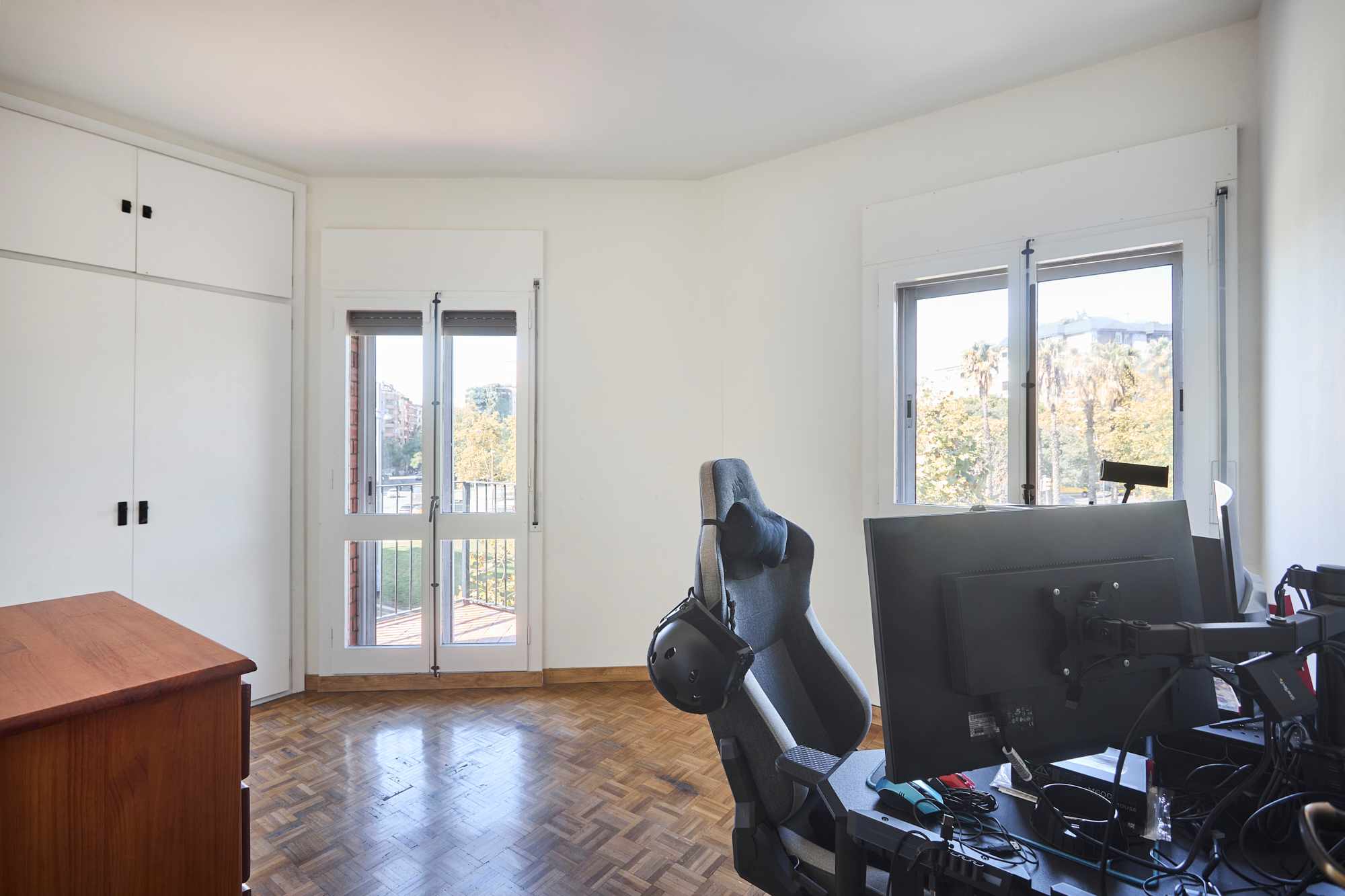 Home office with white cabinets along left wall, wooden desk, and a high-backed chair by a multi-monitor setup near large windows showing trees outside.