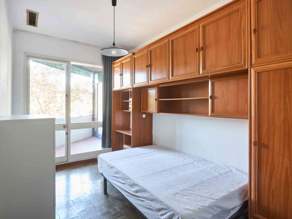 Bedroom with tall wooden cabinets along one wall and a bed at the bottom right, near a glass door letting in light.