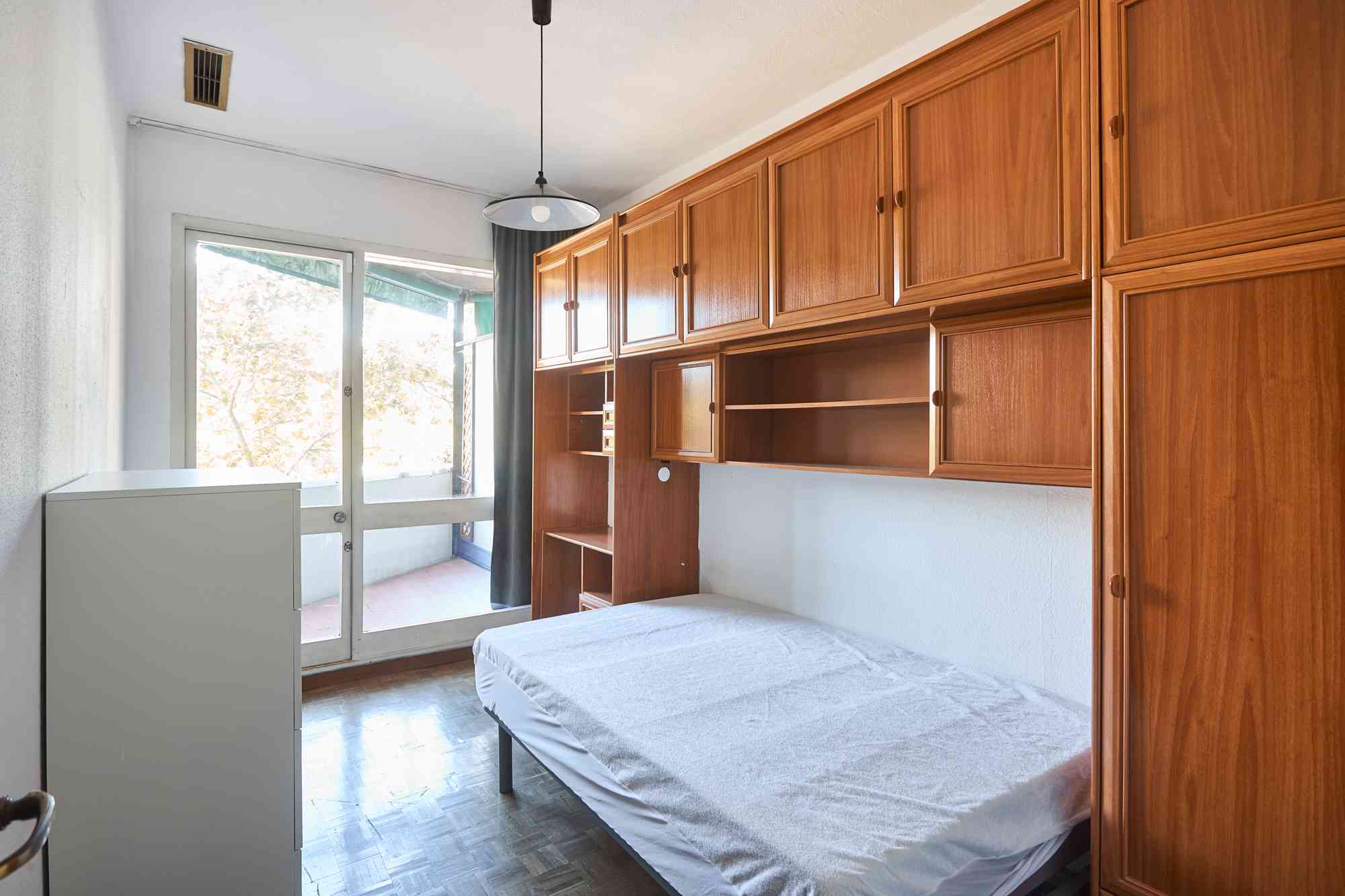 Bedroom with tall wooden cabinets along one wall and a bed at the bottom right, near a glass door letting in light.