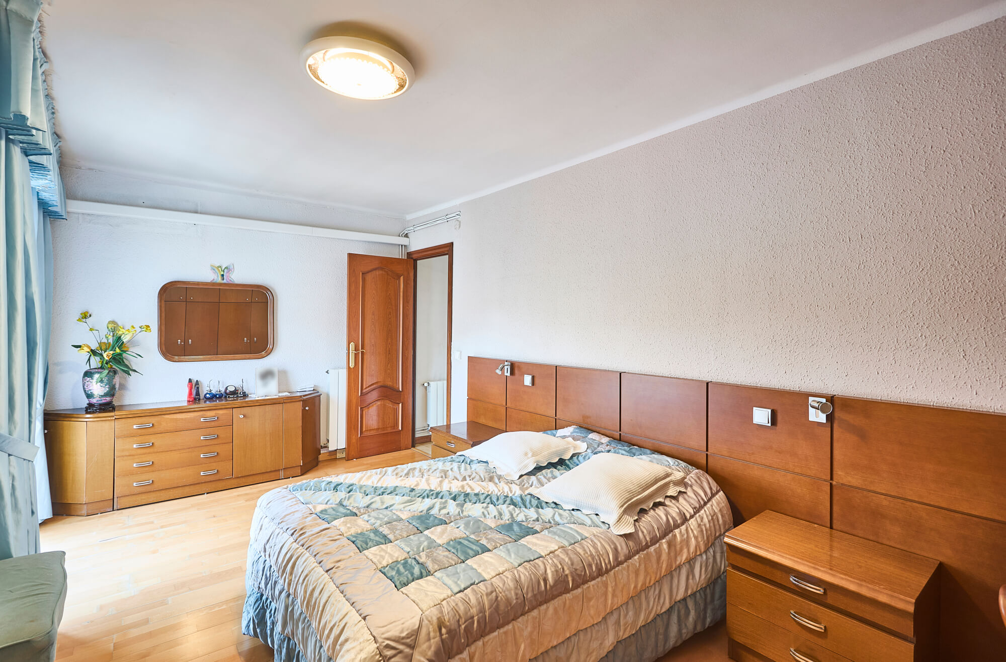 A bright bedroom with two parallel wooden beds, matching dressers, and a wooden door near a built-in wardrobe; blue curtains on the left.