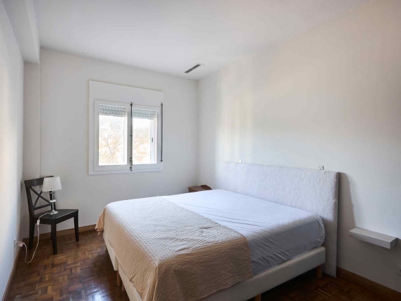 Bright minimalist bedroom with a large bed, white headboard, beige quilt, window with blinds, and wooden parquet floor.