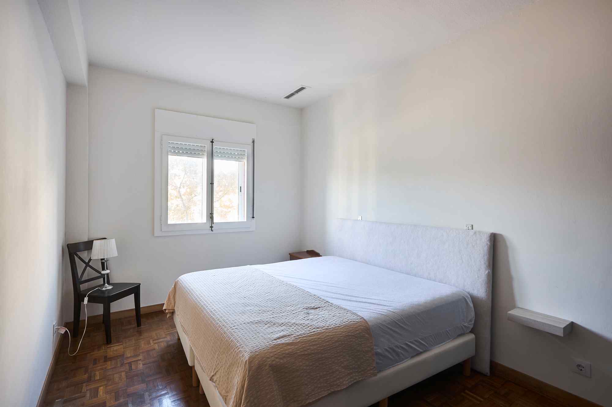 Minimalist bedroom with a large white bed in the foreground, white walls, and a window on the far wall letting in daylight; a chair with a lamp sits to the left.
