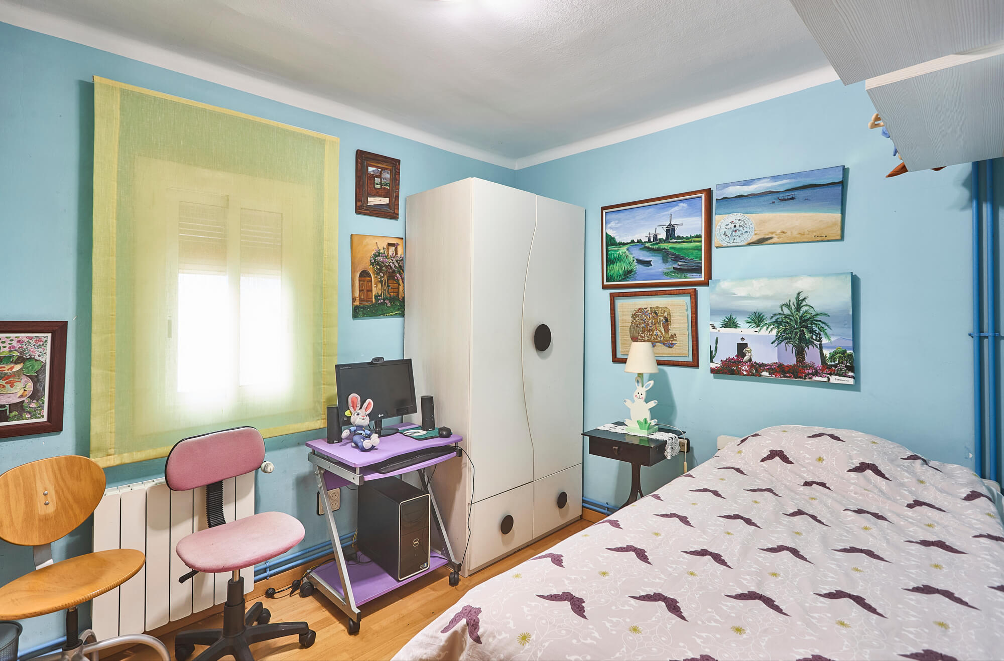 Bedroom with blue walls, a white wardrobe, and a desk area containing a computer setup; bed with a purple-patterned duvet is visible in the foreground.