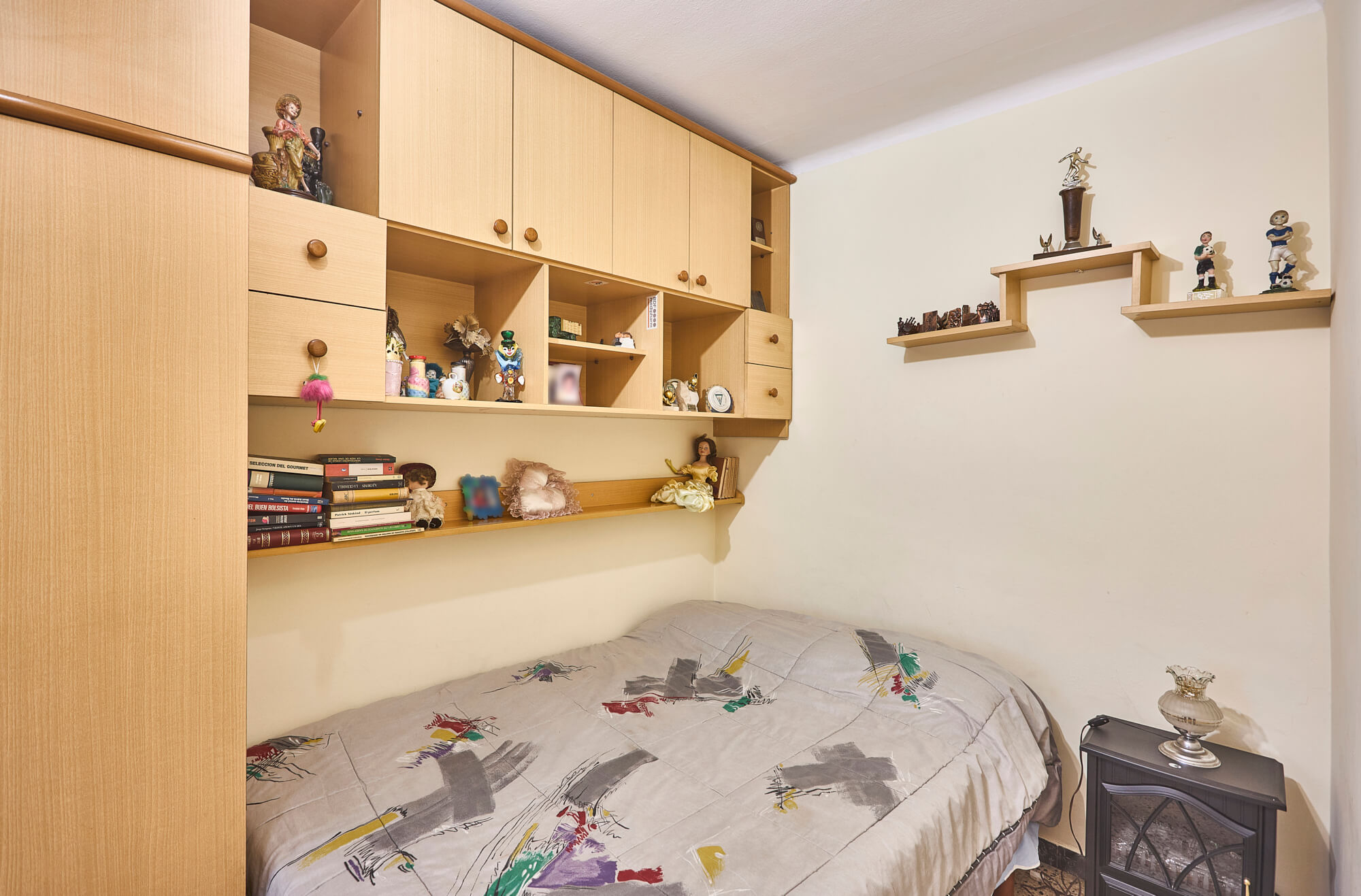 Bedroom with wall-to-wall light-wood cabinets and open shelves filled with figurines and books above a bed.