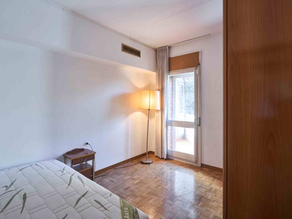 Simple bedroom with a bed in the foreground, small bedside table, and a tall floor lamp near a window with beige curtains; a wooden wardrobe is on the right.