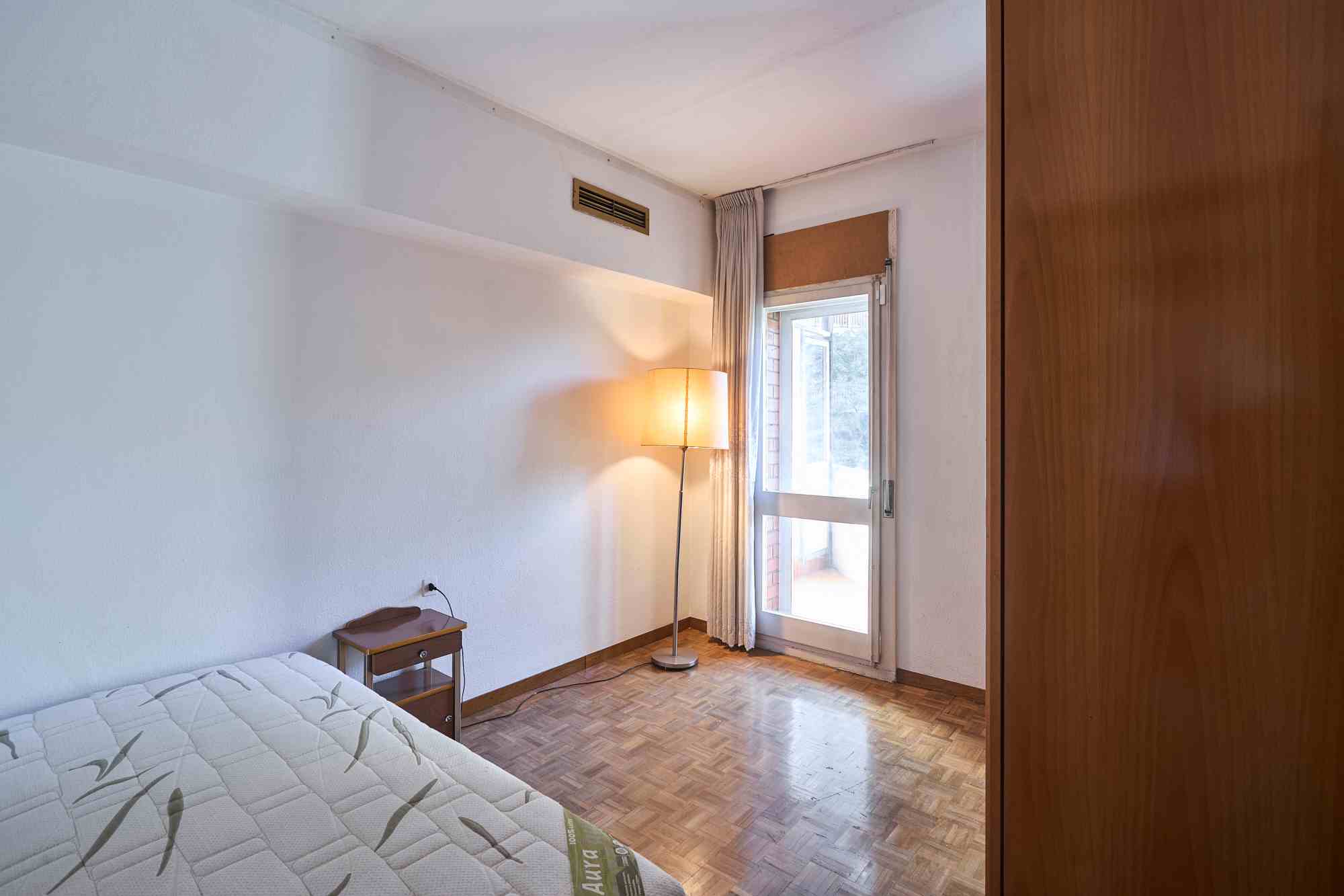 Simple bedroom with a bed in the foreground, small bedside table, and a tall floor lamp near a window with beige curtains; a wooden wardrobe is on the right.