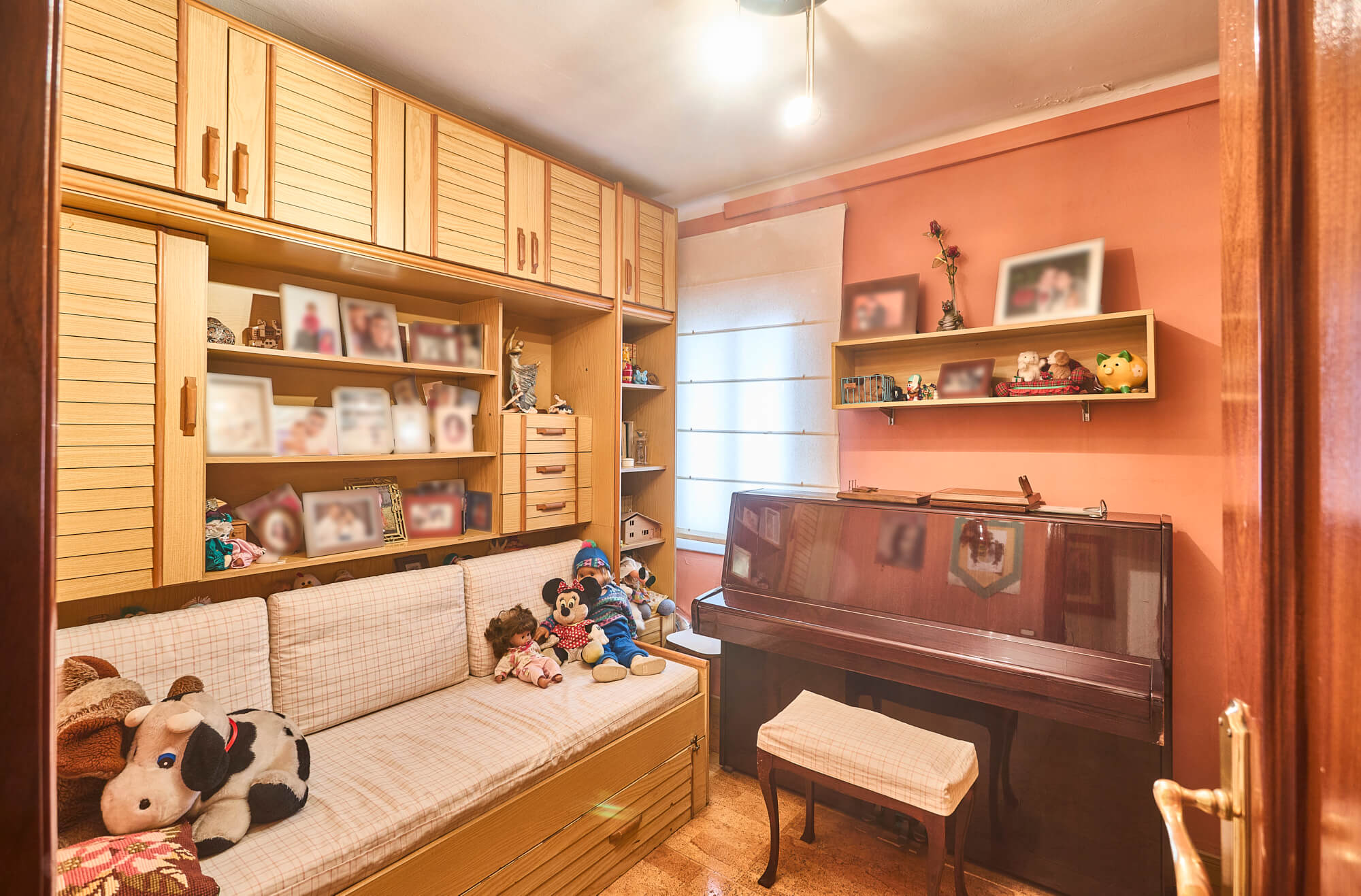 Cozy living room with built‑in wooden cabinets, shelves of family photos, and a piano against pink walls with a plaid sofa and toys nearby.