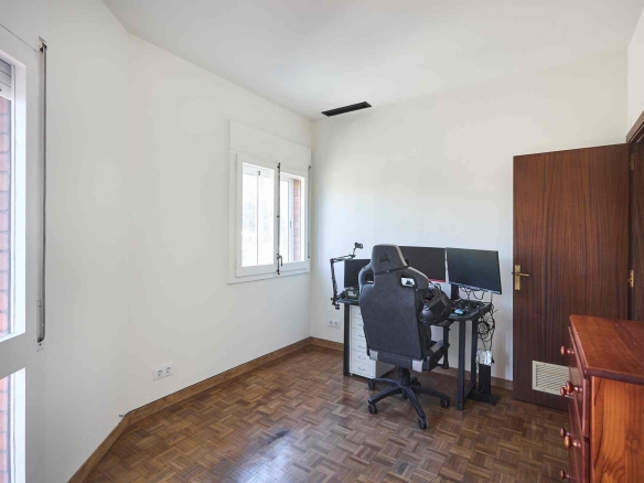 Home office with a desk, dual monitors, and an ergonomic chair by a white wall and windows; wooden floor.