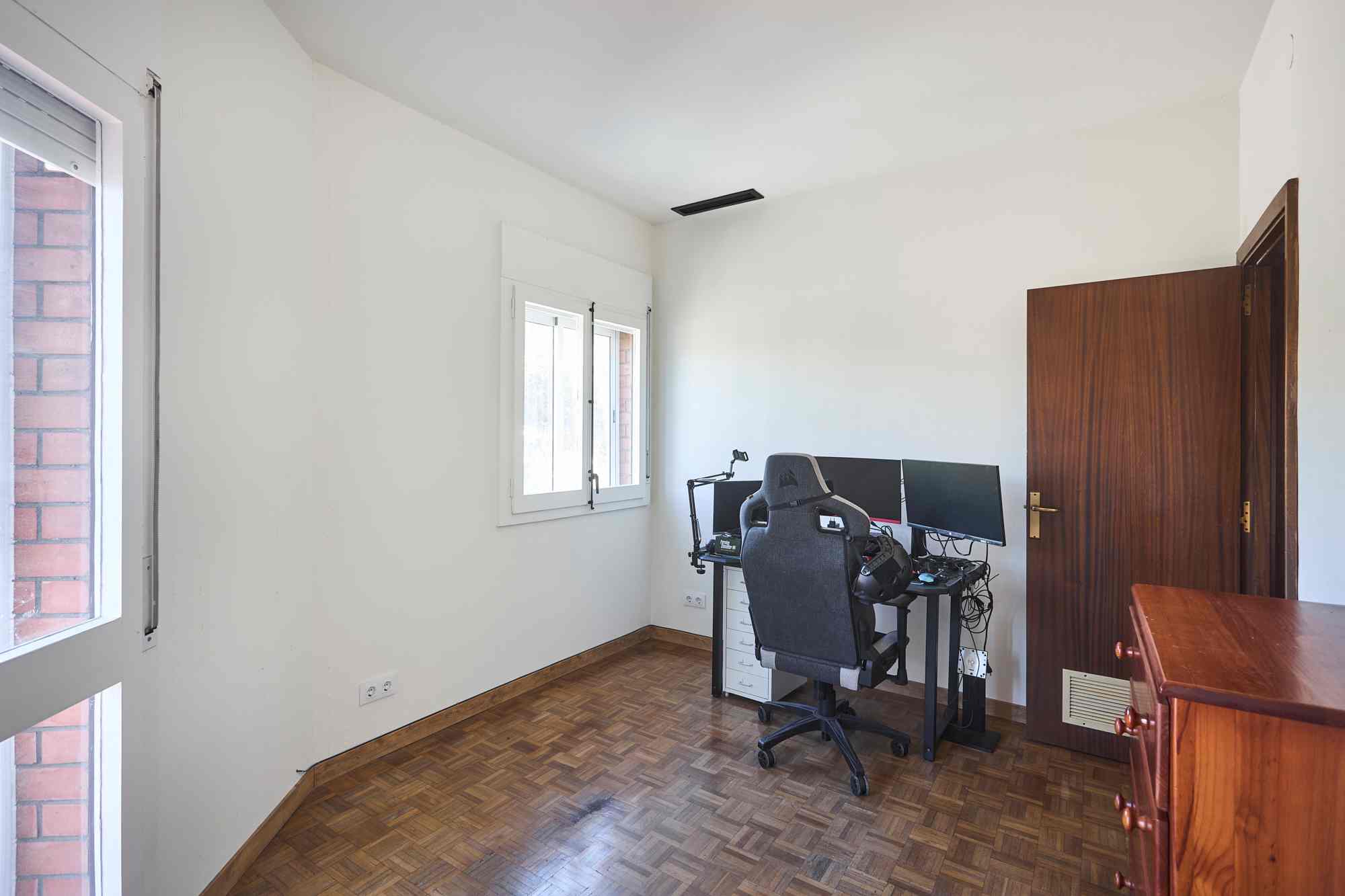 Home office with a desk, dual monitors, and an ergonomic chair by a white wall and windows; wooden floor.