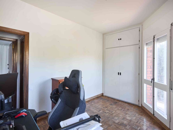 Home office with a gray ergonomic chair, desk cluttered with papers and a monitor, light wooden flooring, and built-in white cabinets next to a glass door to the outside.