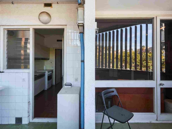 Doorway to a small kitchen with a stove and white tiled walls; a blue downspout runs beside a large window with vertical blinds showing outdoor greenery; a folding chair on the tiled floor.