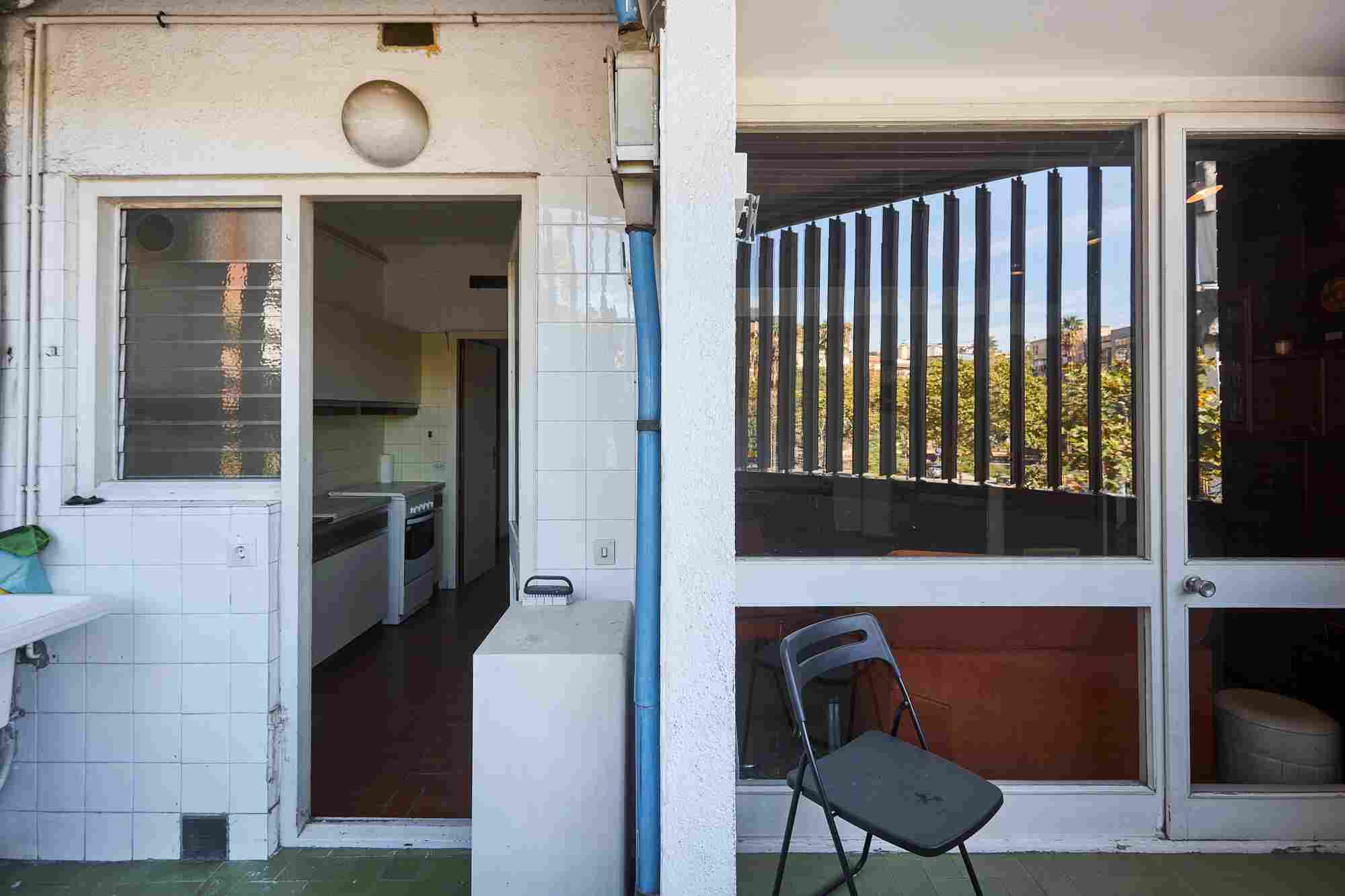 Doorway to a small kitchen with a stove and white tiled walls; a blue downspout runs beside a large window with vertical blinds showing outdoor greenery; a folding chair on the tiled floor.