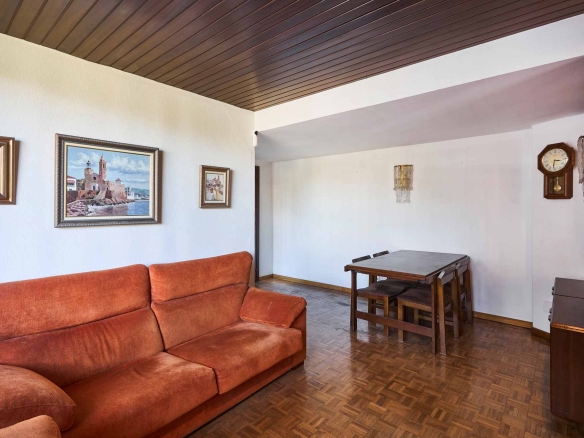 Living room with an orange sectional sofa, wooden parquet floor, white walls, and framed landscape paintings; a dining table is in the background.