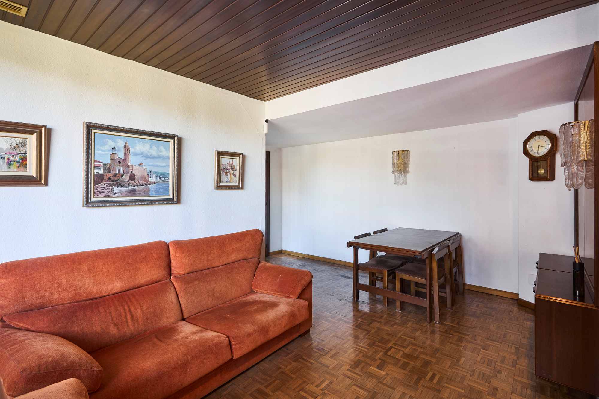 Living room with an orange sectional sofa, wooden parquet floor, white walls, and framed landscape paintings; a dining table is in the background.