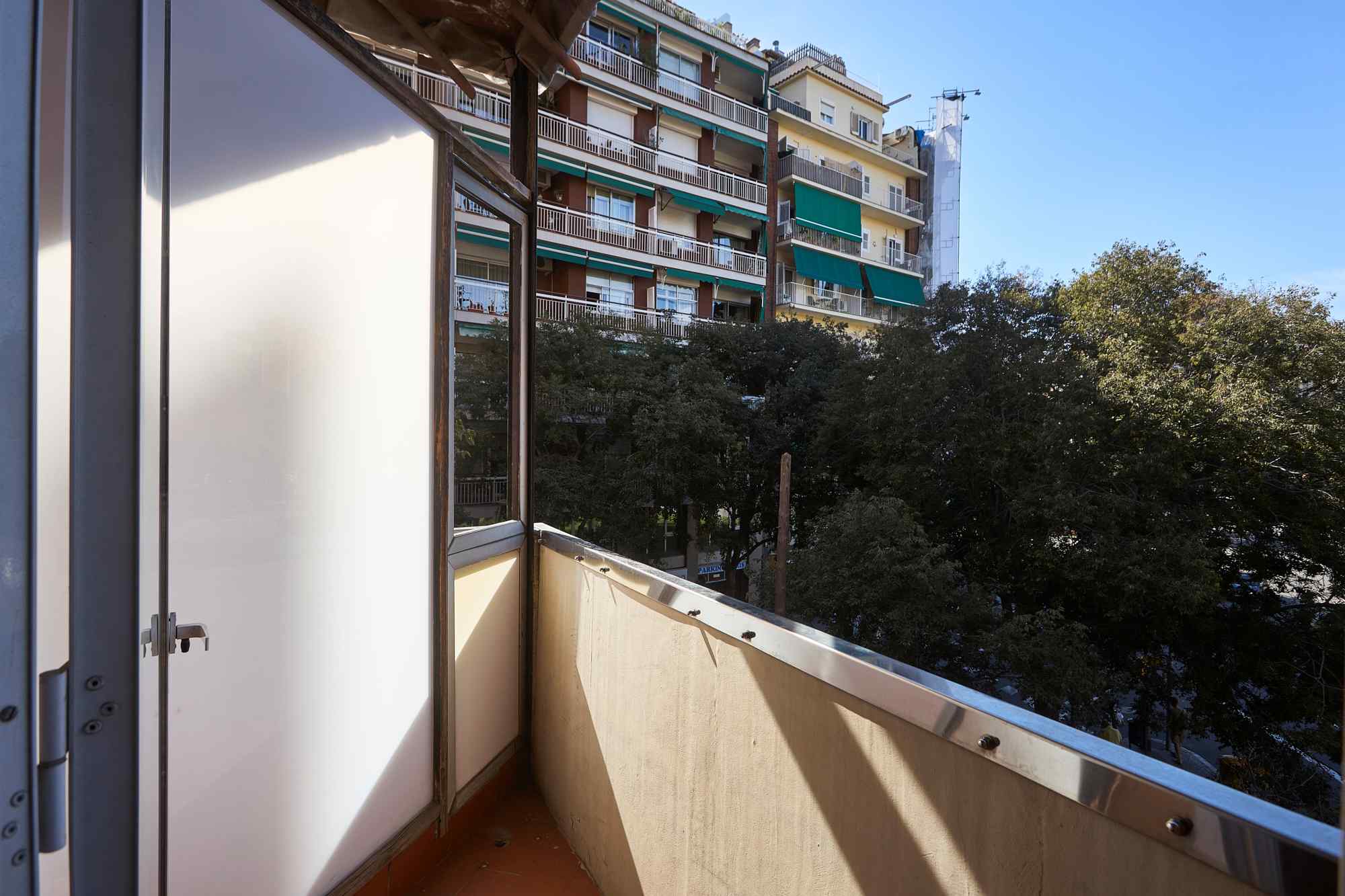 Balconies of an urban apartment building with green awnings seen from a small balcony and trees below, blue sky above.