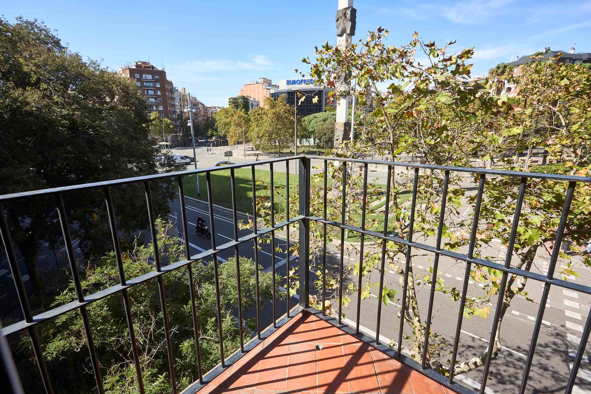 View from a balcony overlooking a tree-lined city street with mid-rise buildings and a clear blue sky.