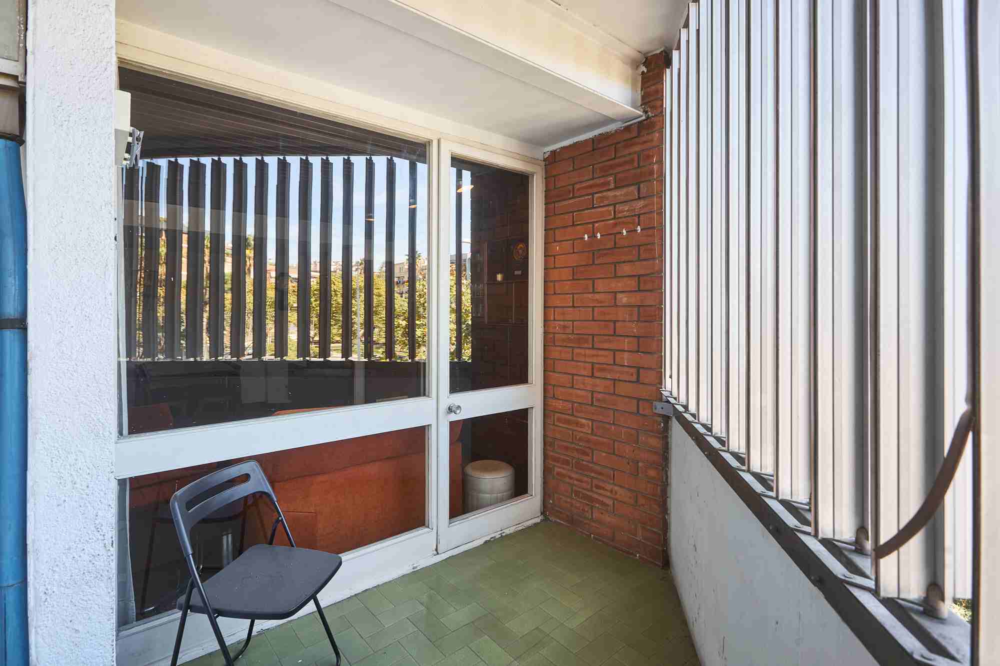 Balcony area with sliding glass doors, brick wall, and a black folding chair on a green tiled floor.